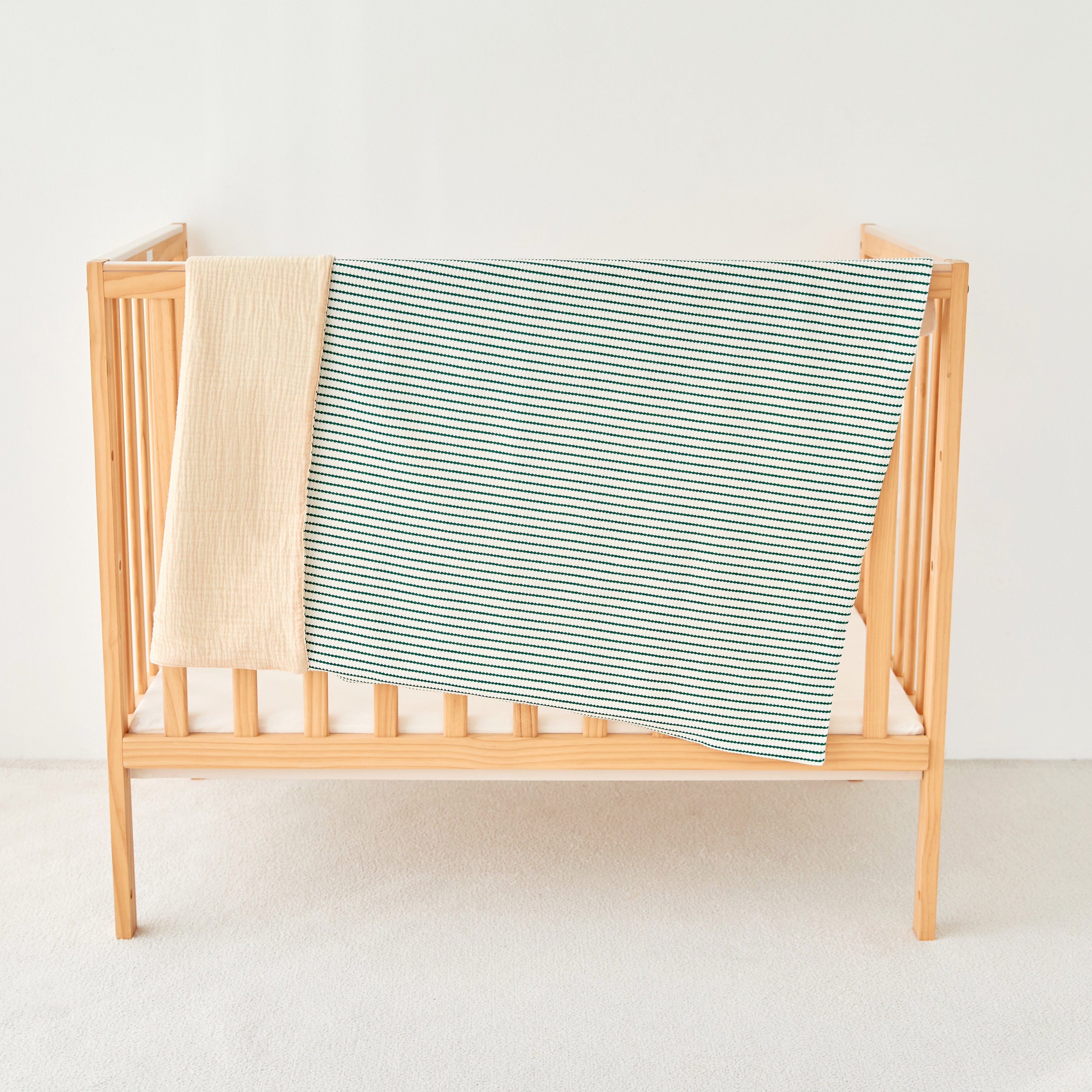 Wooden baby crib with a green and white striped knitted gauze blanket draped over it on a white background