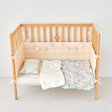 Wooden crib with beige bumper pad, white blanket, and baby zoo animal patterned quilt on a light gray floor.