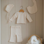 Baby small floral pattern pajama set with hat, bib, and pants hanging on a wooden shelf against a beige paneled wall.