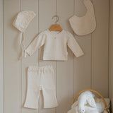 Baby small floral pattern pajama set with hat, bib, and pants hanging on a wooden shelf against a beige paneled wall.
