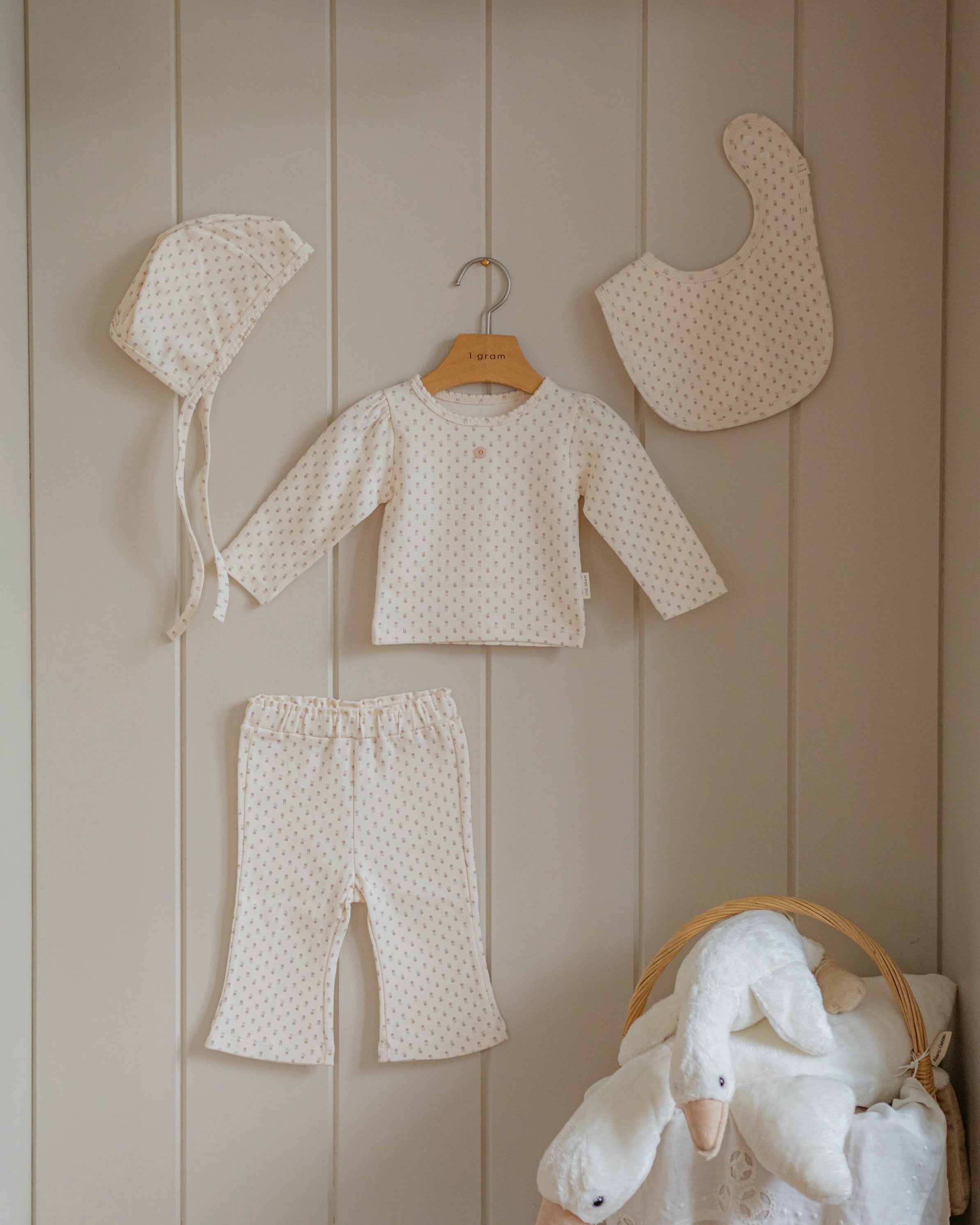 Baby small floral pattern pajama set with hat, bib, and pants hanging on a wooden shelf against a beige paneled wall.