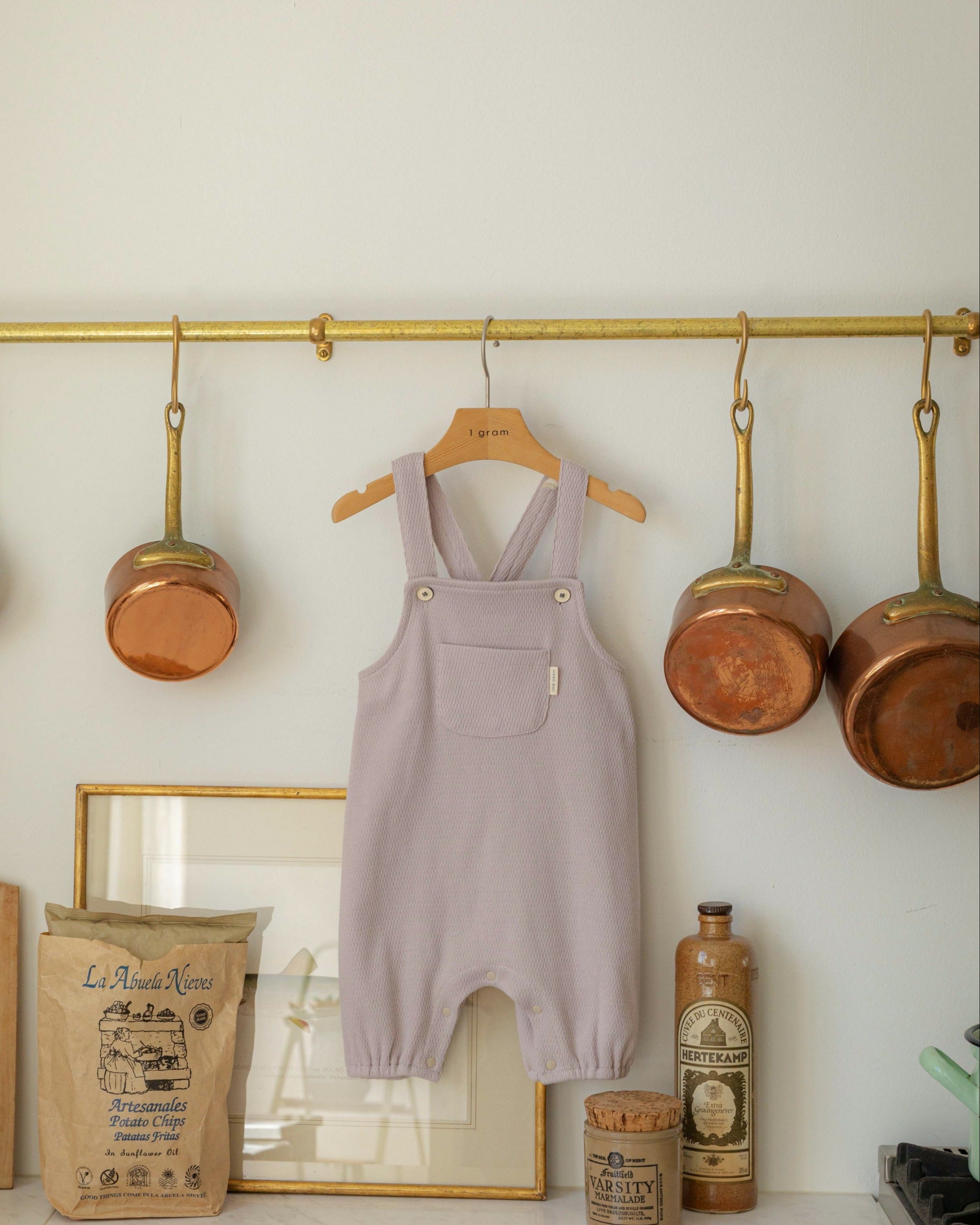 Purple children's overalls hanging on a rack in a kitchen with pots and a framed picture.