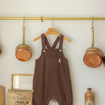 Brown children's overalls hanging on a gold hook against a white wall with decorative items.