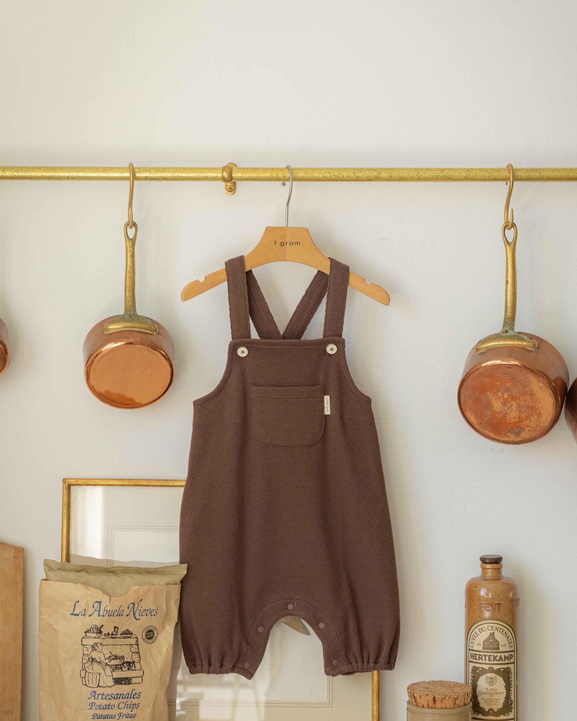 Brown children's overalls hanging on a gold hook against a white wall with decorative items.