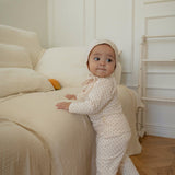 Baby in a small floral pattern ivory outfit standing on a couch in a room with wooden flooring and white walls.
