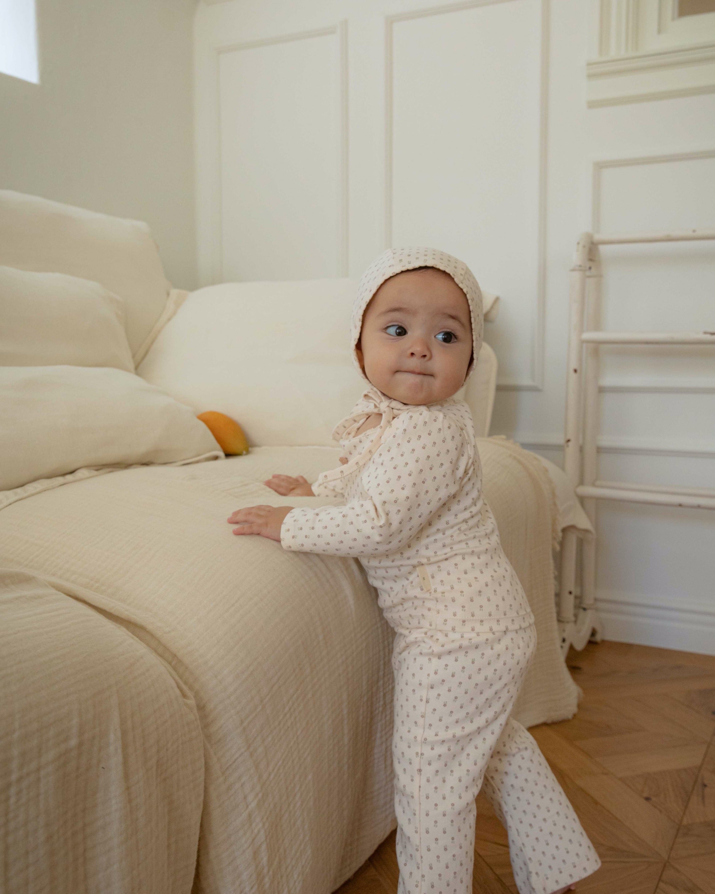 Baby in a small floral pattern ivory outfit standing on a couch in a room with wooden flooring and white walls.