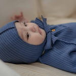 Baby wearing a blue romper onesie with a bonnet on a light-colored surface