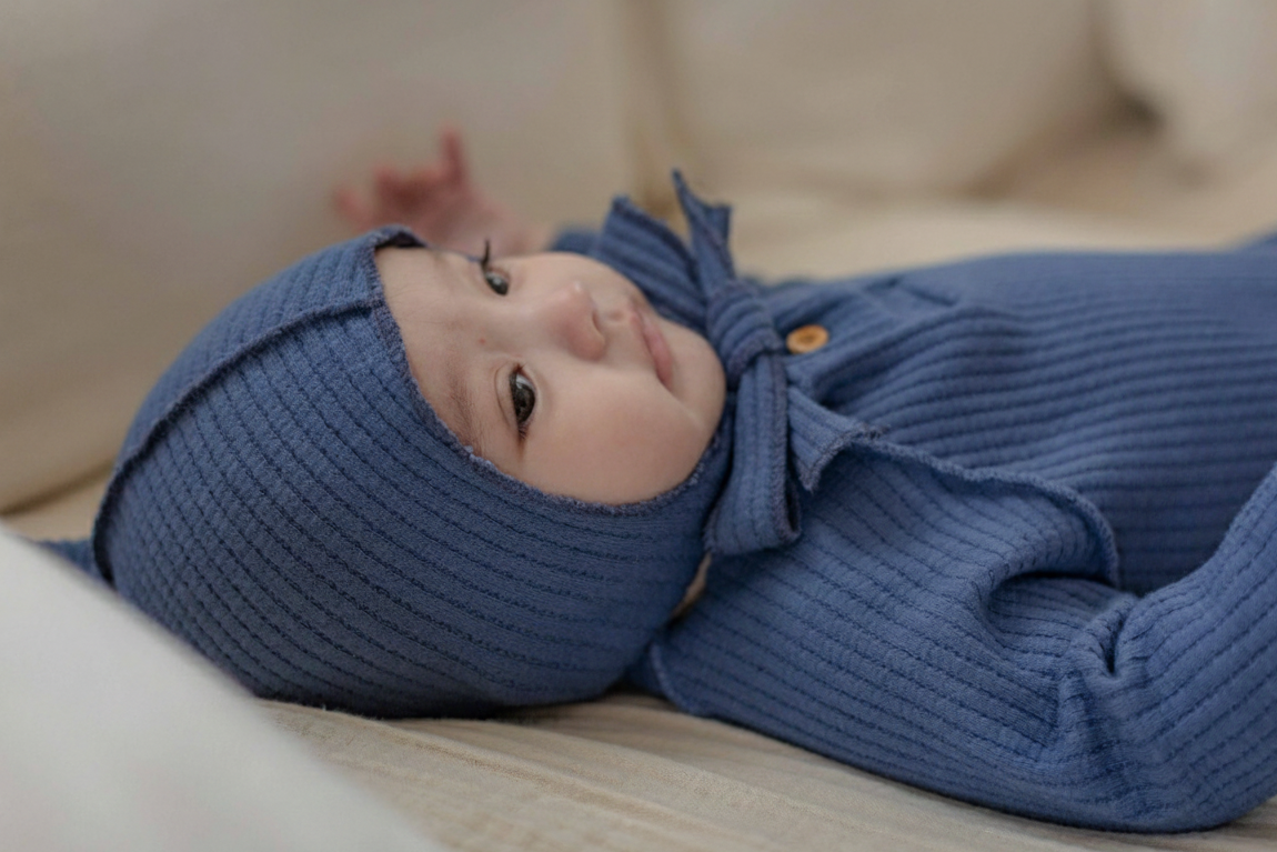 Baby wearing a blue romper onesie with a bonnet on a light-colored surface