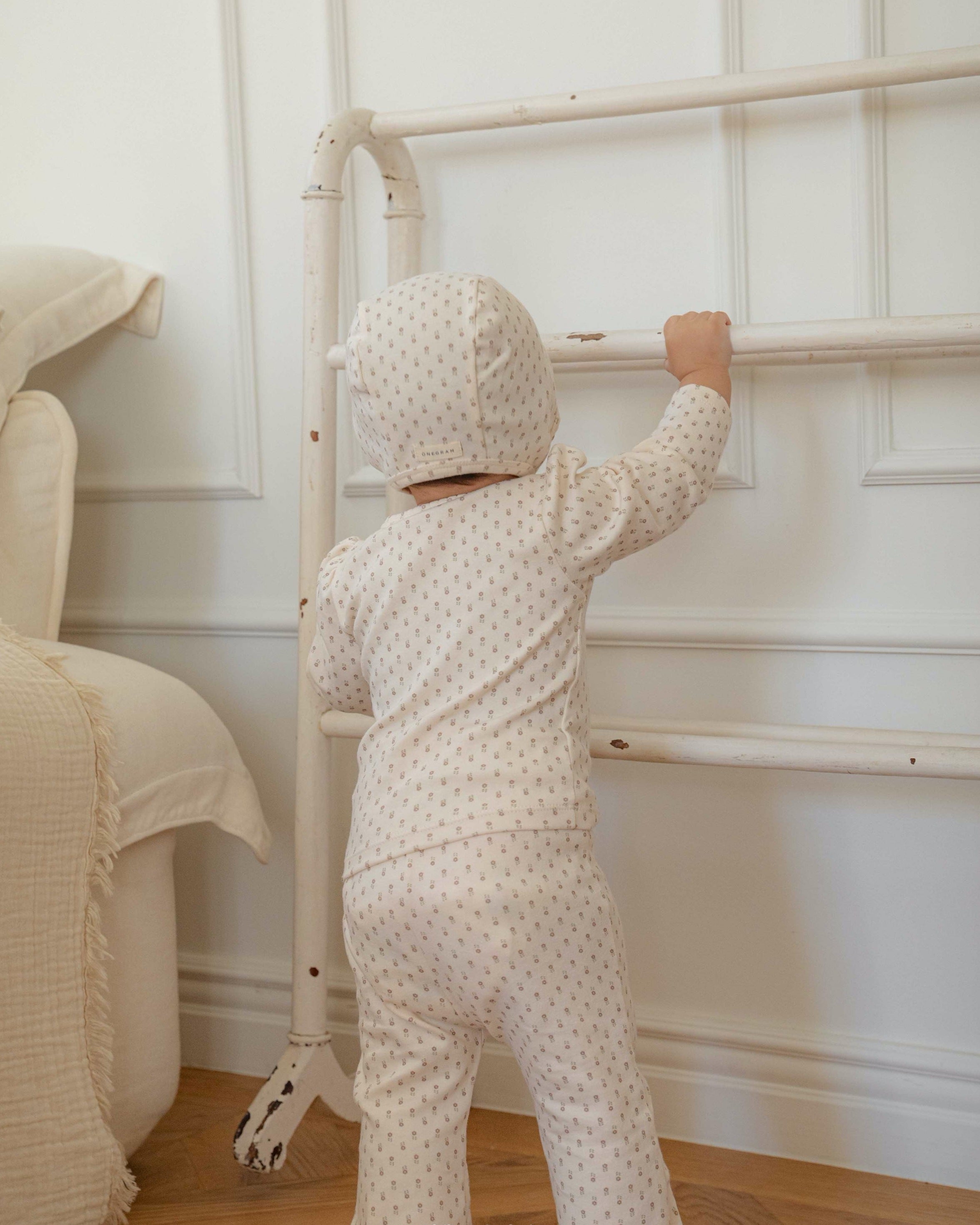 Child in a small floral print outfit standing next to a crib in a nursery.