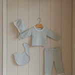 Baby outfit set with pajama long sleeves pants, bib, and hat hanging on a wooden shelf.