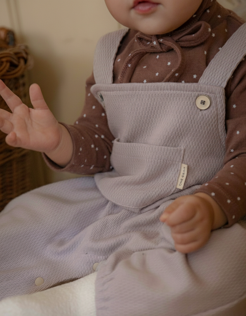 Baby wearing a brown polka dot shirt and light purple overalls sitting on a white surface.