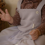 Baby wearing a brown polka dot shirt and light purple overalls sitting on a white surface.