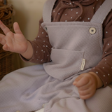 Baby wearing a brown polka dot shirt and light purple overalls sitting on a white surface.