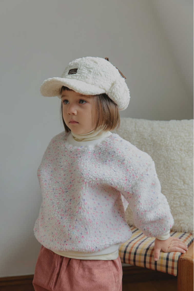 Child wearing a textured winter white pink sweater and beige cap sitting on a plaid cushion.