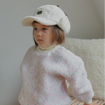 Child wearing a textured winter white pink sweater and beige cap sitting on a plaid cushion.