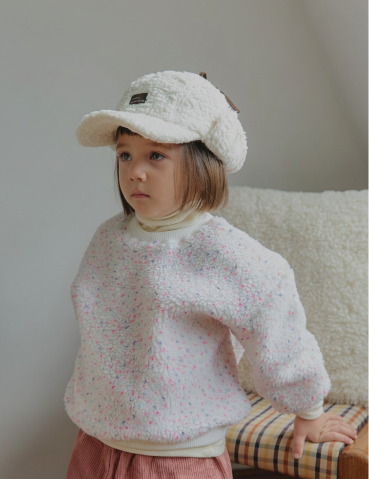 Child wearing a textured winter white pink sweater and beige cap sitting on a plaid cushion.