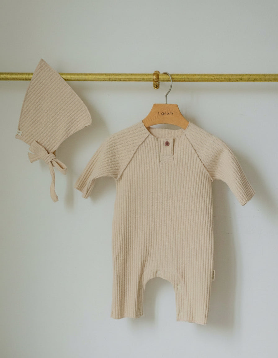 Beige baby romper and bonnet hanging on a gold hook against a white wall.