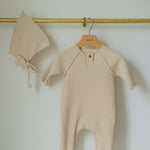 Beige baby romper and bonnet hanging on a gold hook against a white wall.