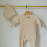 Beige baby romper and bonnet hanging on a gold hook against a white wall.