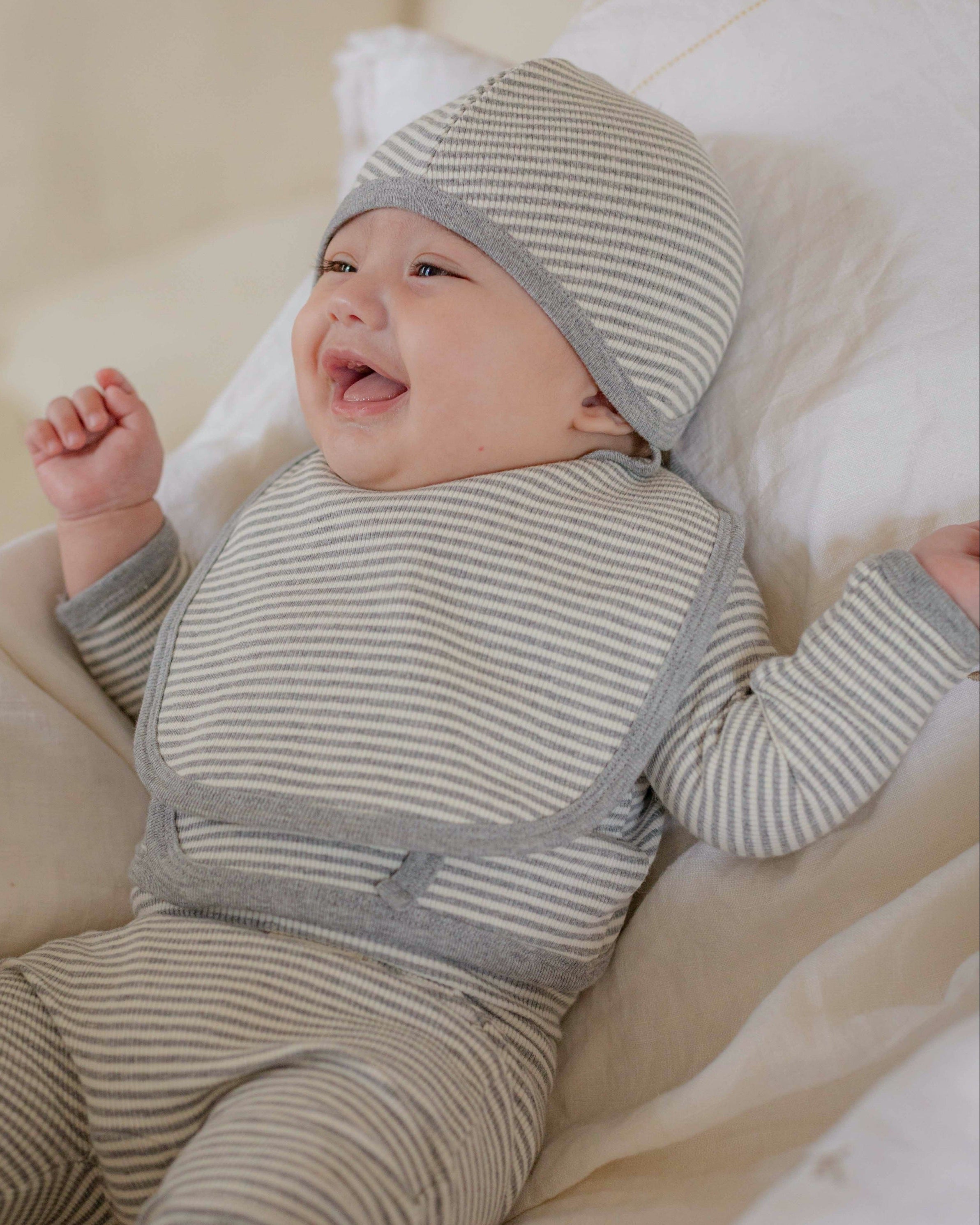 Baby in a gray white striped onesie outfit and hat lying on a soft surface