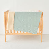 Wooden baby crib with a green and white striped knitted gauze blanket draped over it on a white background