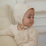 Baby wearing a cream-colored bonnet and outfit with small flower pattern, standing by a couch.