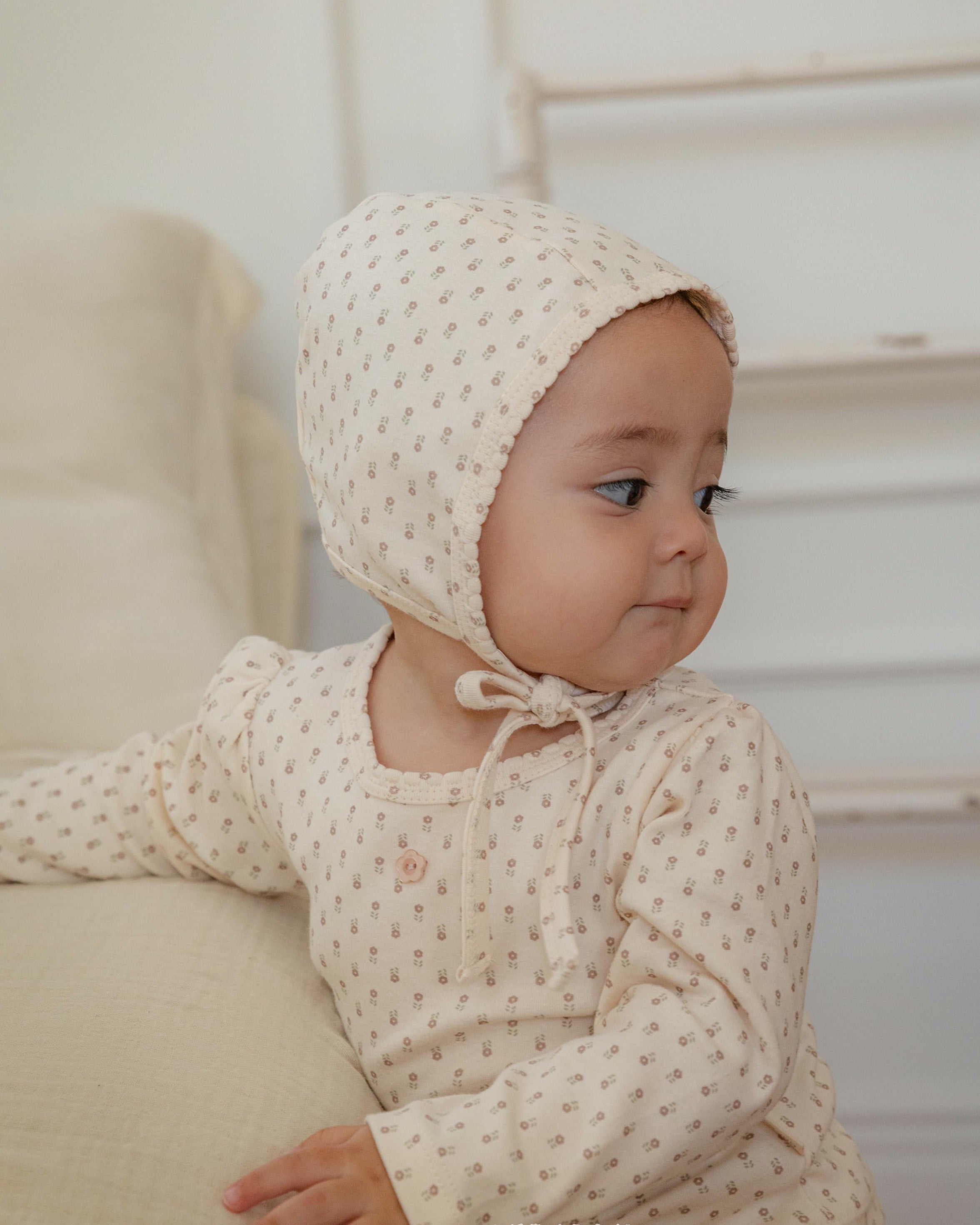 Baby wearing a cream-colored bonnet and outfit with small flower pattern, standing by a couch.