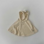 Beige baby cape dress with pompom attached hood on a light gray background