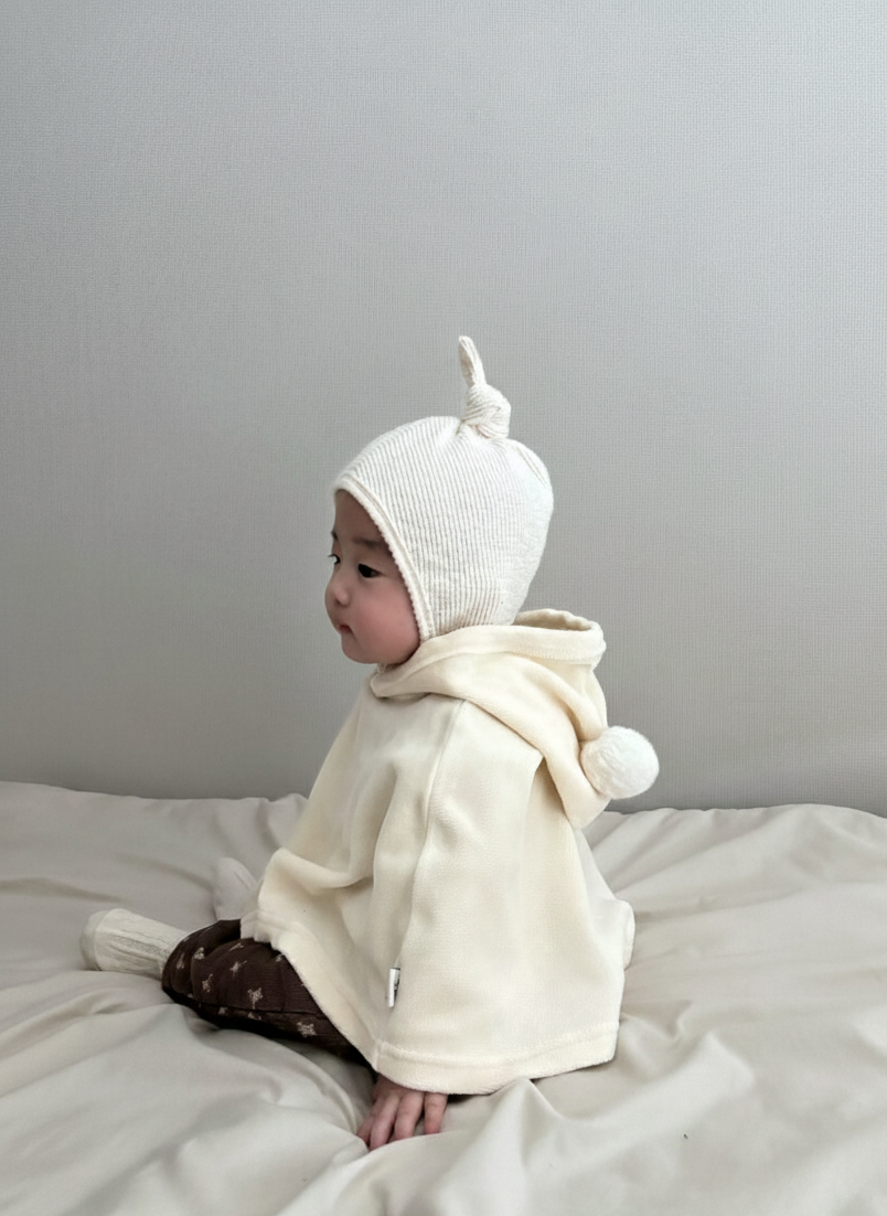 Baby wearing a white knit hat and cream-colored hoodie sitting on a light gray surface.