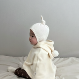Baby wearing a white knit hat and cream-colored hoodie sitting on a light gray surface.