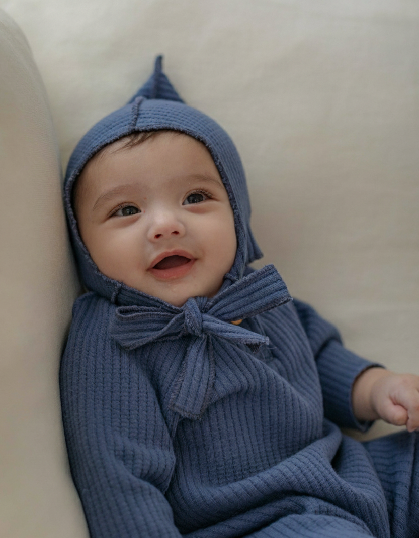 Baby wearing navy blue knitted outfit with a hood and bow tie, sitting on a beige surface.