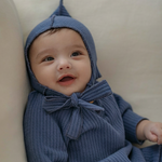 Baby wearing navy blue knitted outfit with a hood and bow tie, sitting on a beige surface.