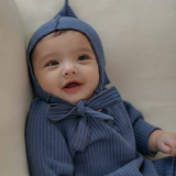 Baby wearing navy blue knitted outfit with a hood and bow tie, sitting on a beige surface.