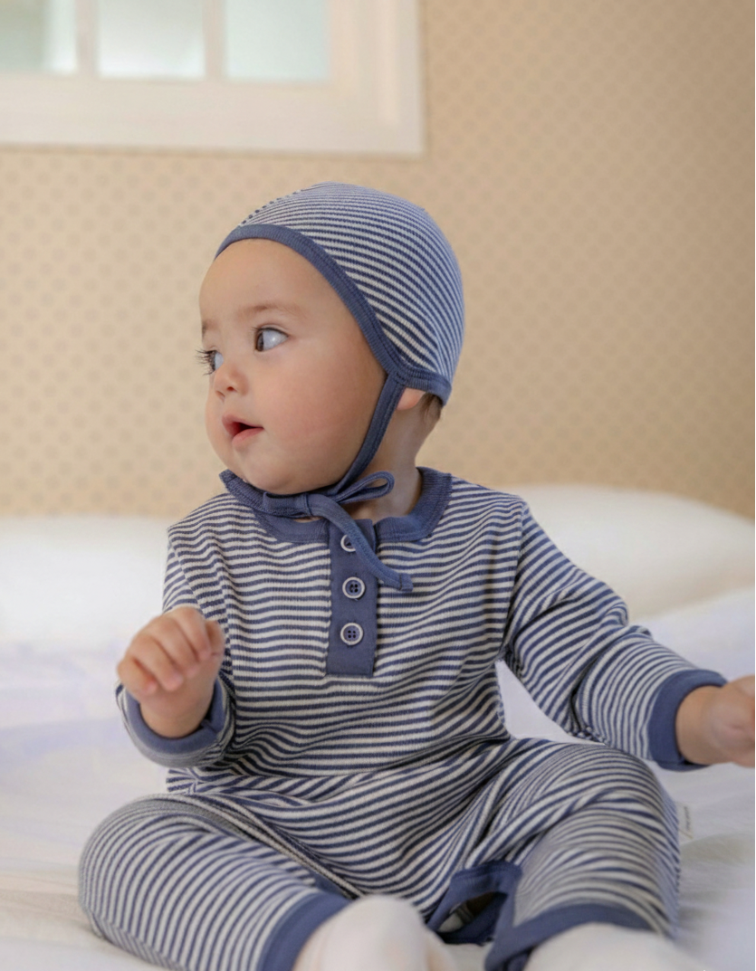 Baby wearing a blue white striped outfit and bonnet sitting on a bed.