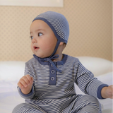 Baby wearing a blue white striped outfit and bonnet sitting on a bed.