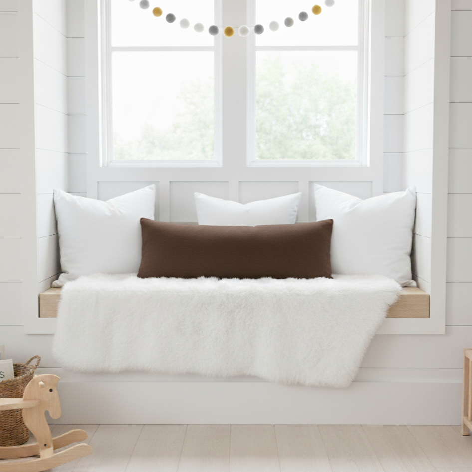 Children's window seat with white cushions and a brown long lumbar pillow, wooden rocking horse, and basket in a bright room.