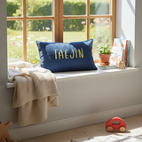 Nursery with a window seat, blue pillow with custom embroidery text, and toys on a wooden floor.