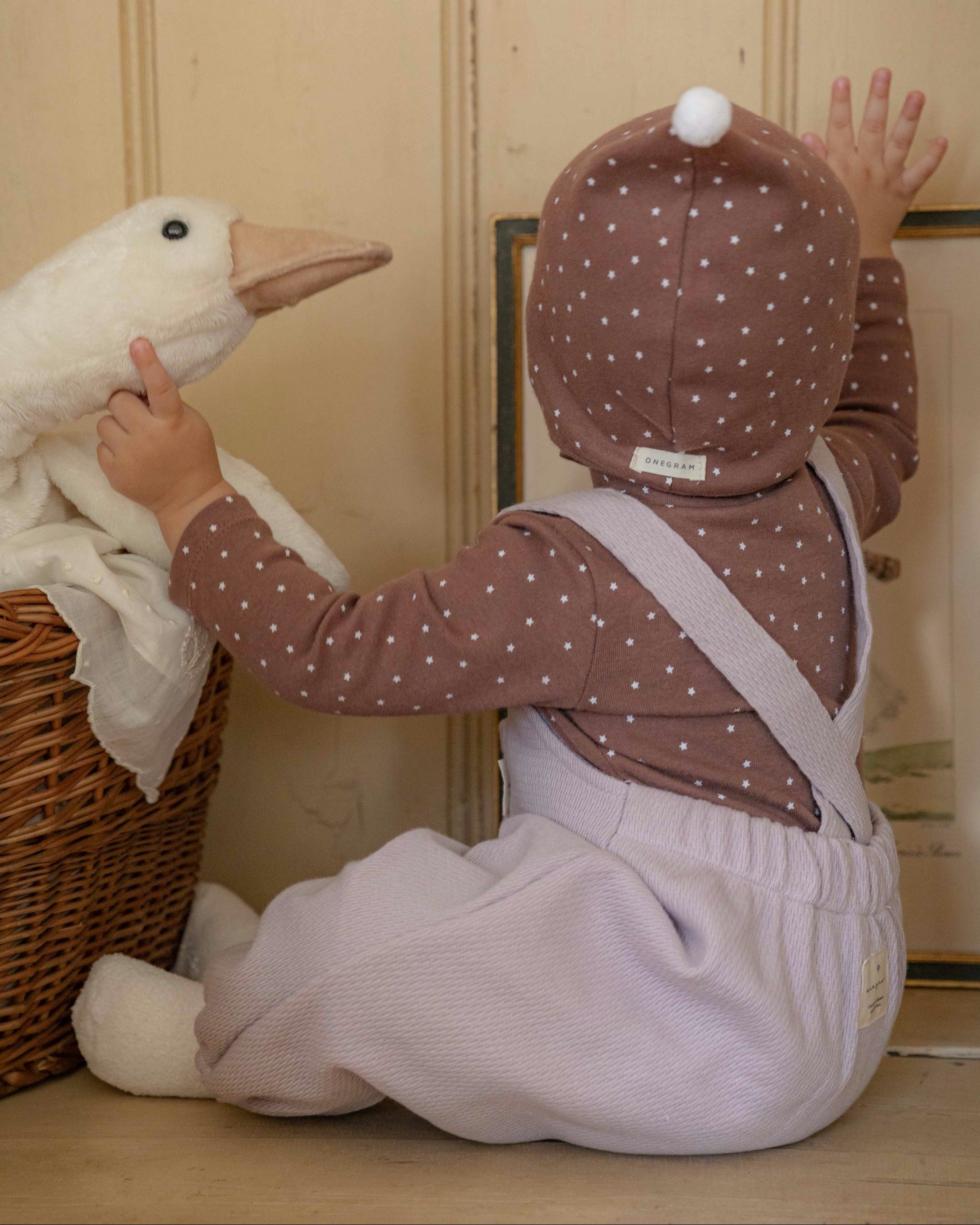 Child in a violet romper, brown outfit with white polka dots playing with a toy goose in a room.