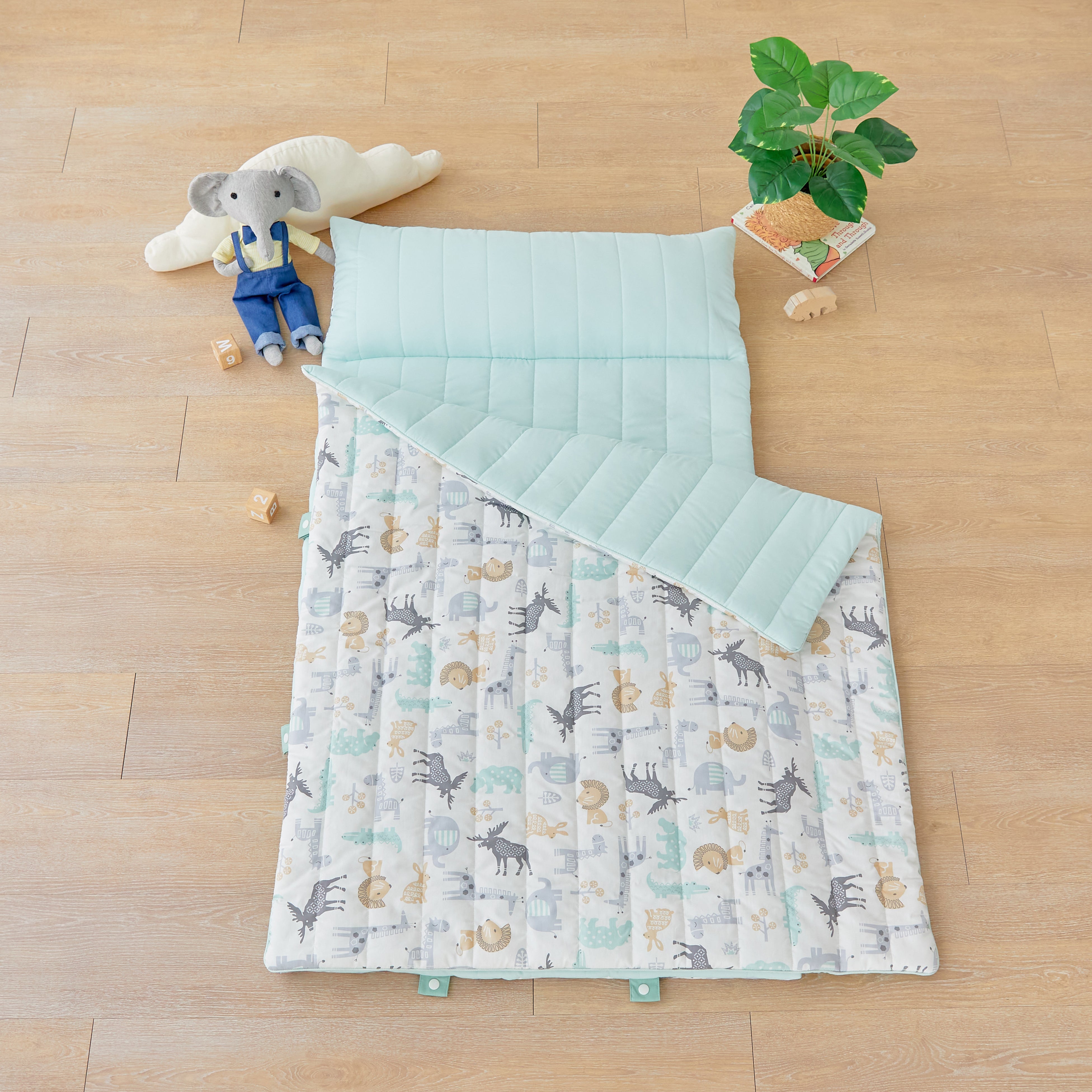 Children's quilt portable nap mat blanket with animal pattern on a wooden floor with toys and a plant.