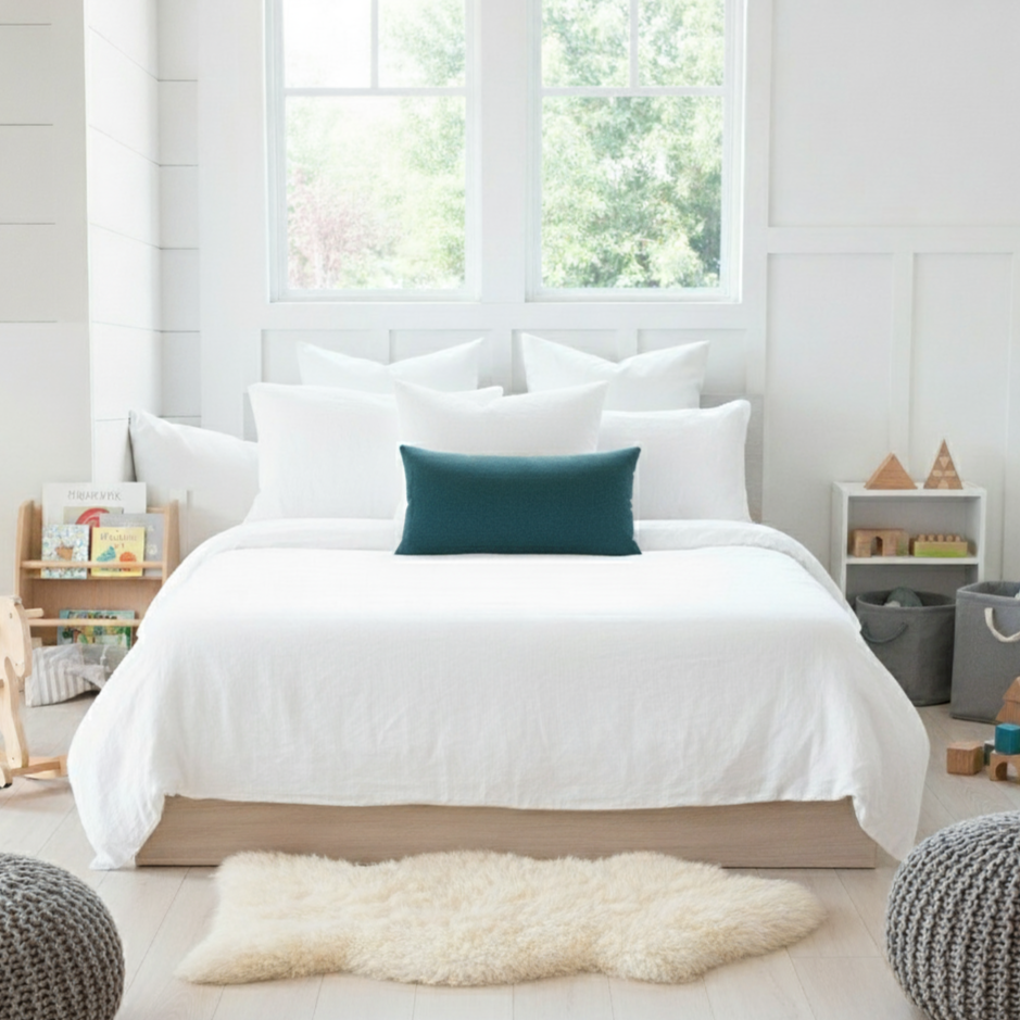 Bedroom with white bedding and teal blue lumbar pillow, light wood floors, and large windows.