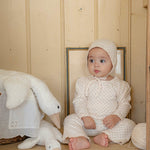 Baby in a white floral outfit sitting on a wooden bench with a plush toy, in a room with wooden walls.