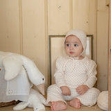 Baby in a white floral outfit sitting on a wooden bench with a plush toy, in a room with wooden walls.