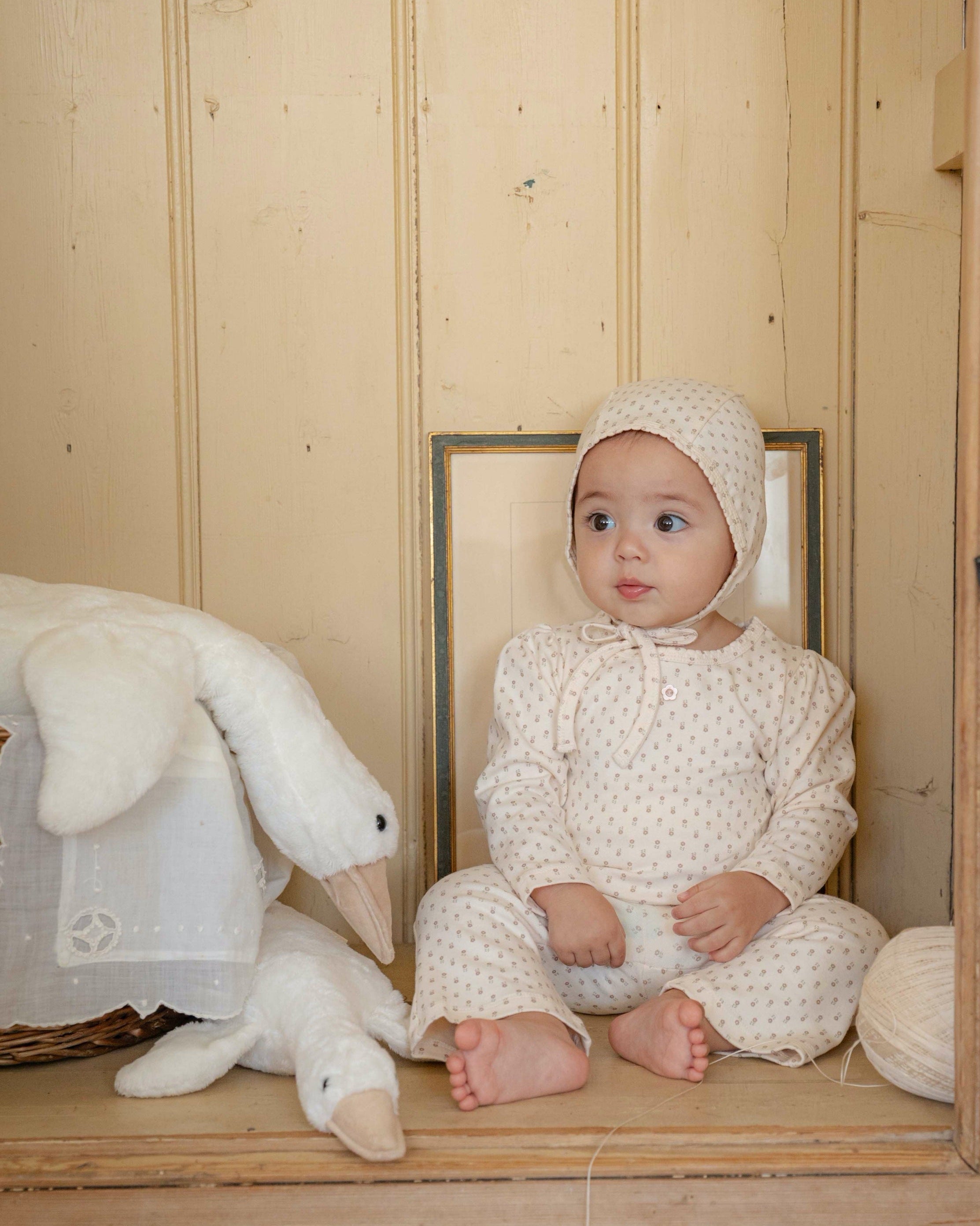 Baby in a white floral outfit sitting on a wooden bench with a plush toy, in a room with wooden walls.