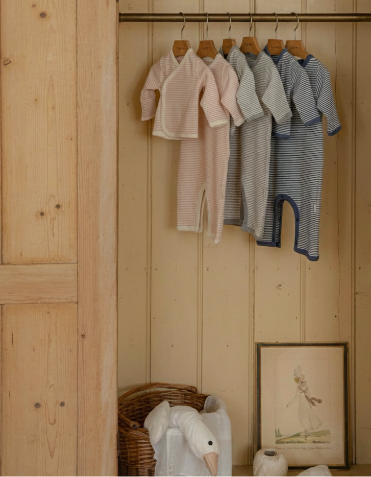 Wooden wardrobe with striped baby onesies clothes in pink, gray, navy blue hanging on a rack inside.