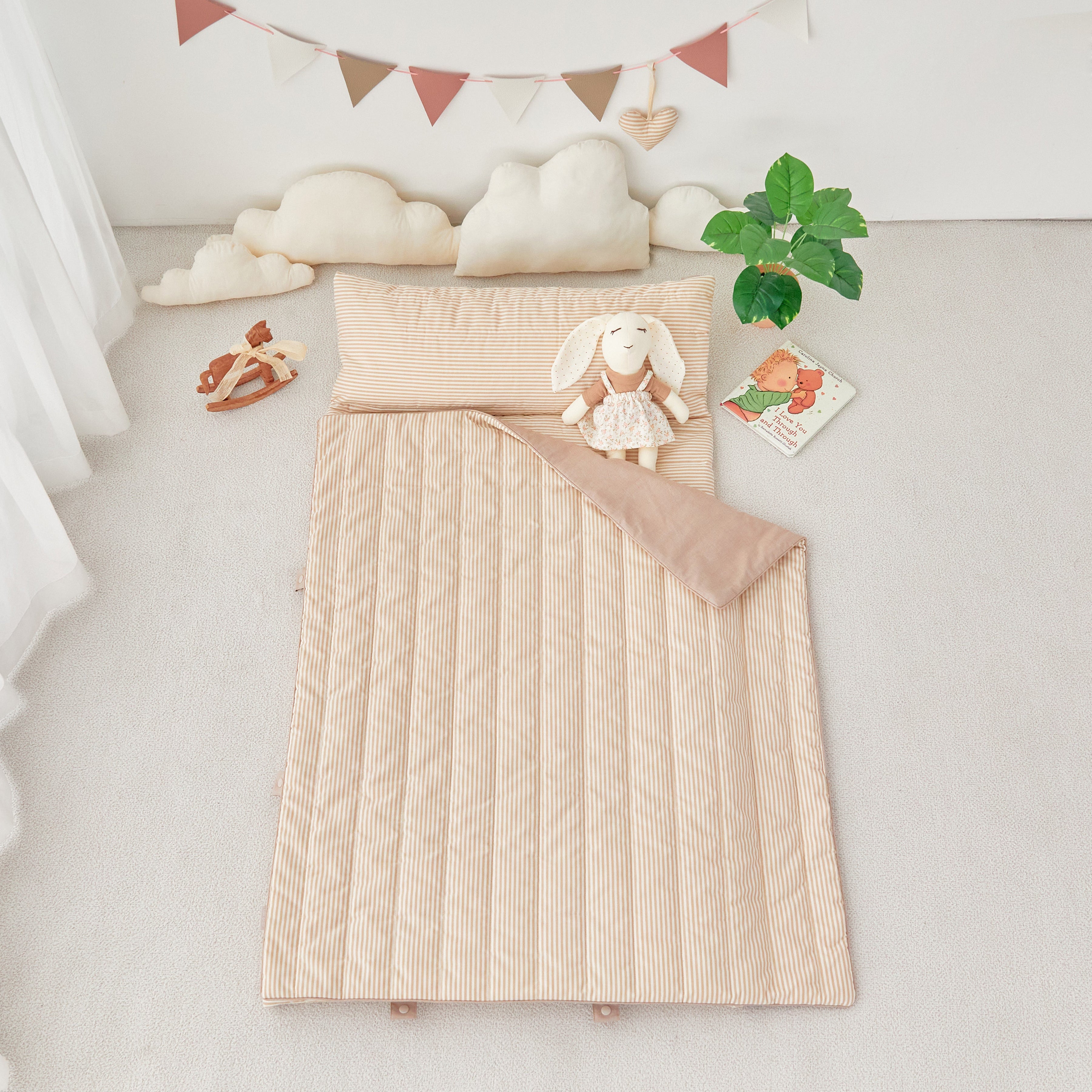 Tan Beige quilted portable nap mat blanket with a white rabbit plush toy on a light gray surface, with decorative elements like clouds and a banner in the background.