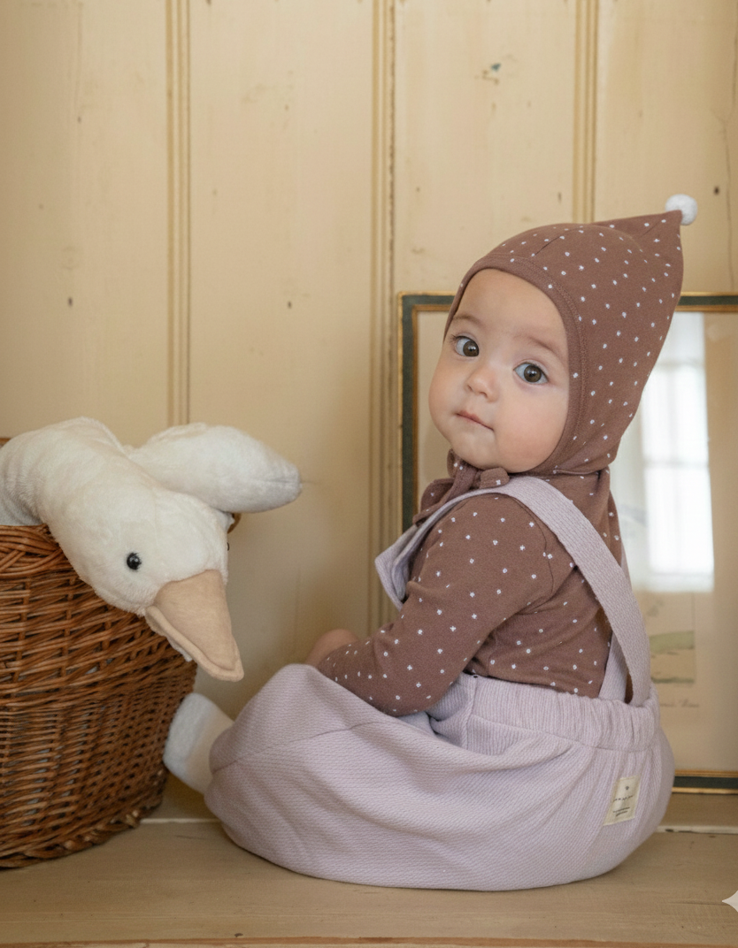 Baby in a brown polka dot outfit and romper overalls with a white plush toy in a wooden room.