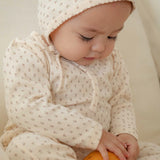 Baby in a white outfit with small floral patterns holding an orange ball on a light background