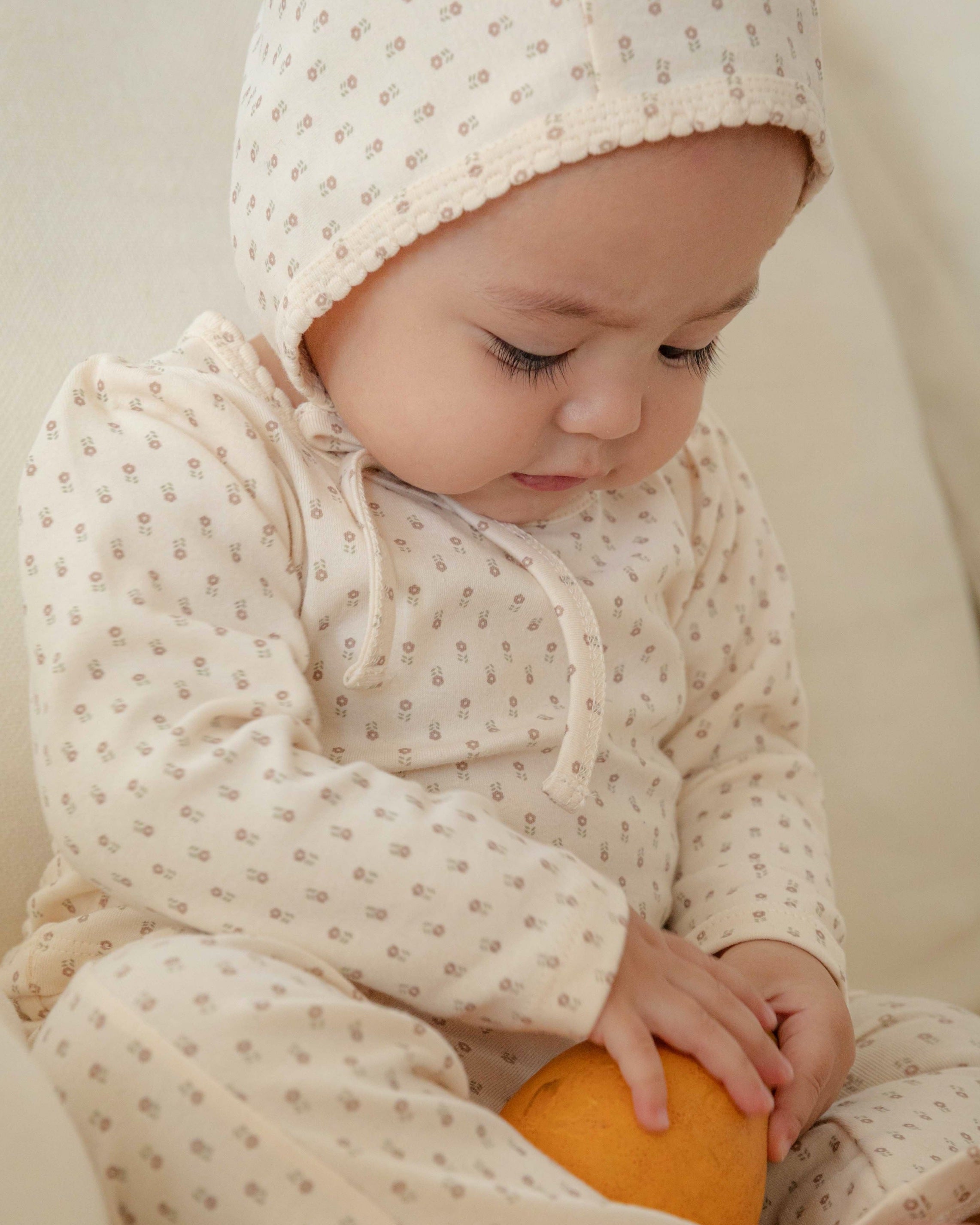 Baby in a white outfit with small floral patterns holding an orange ball on a light background