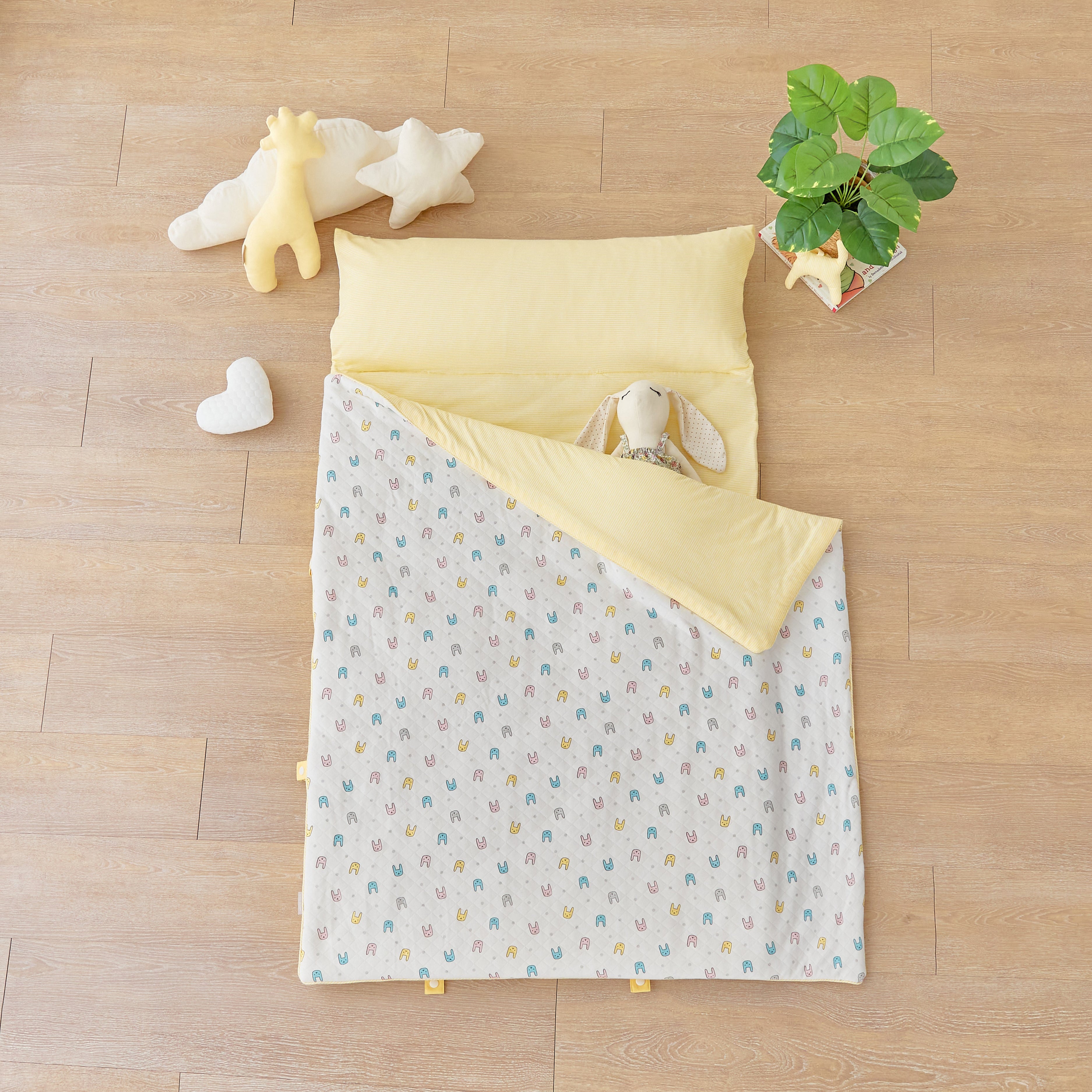 Baby portable nap mat blanket with yellow striped and blue pink bunny rabbit pattern on a wooden floor with toys and a plant.