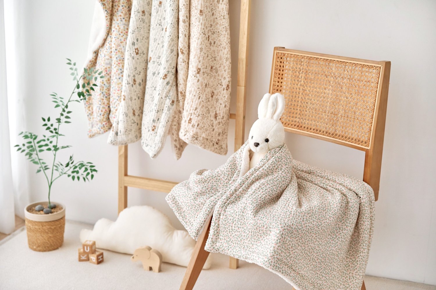 Baby floral gauze blanket with a bunny design draped over a chair in a room with hanging baby clothes and toys.